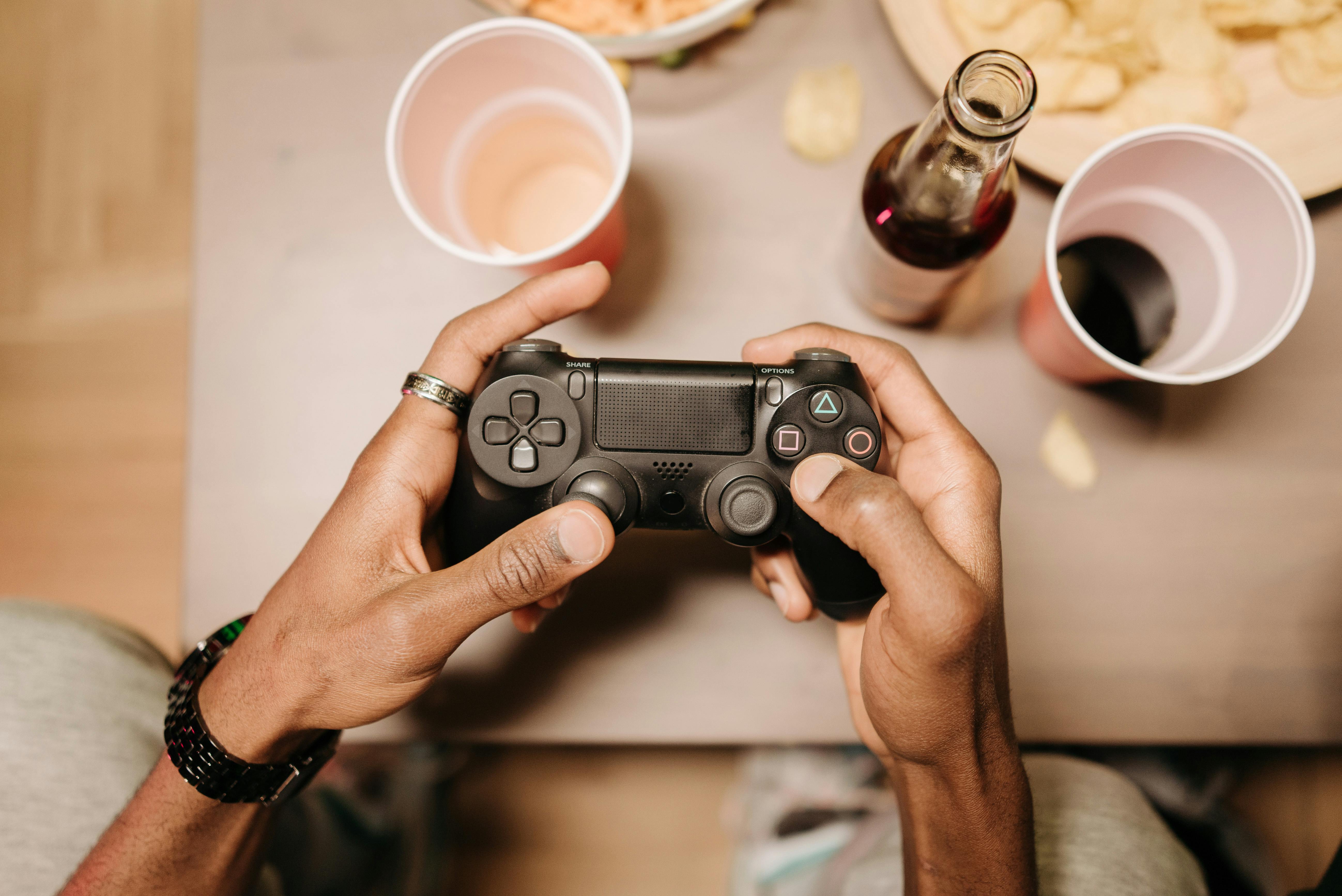 A person holding a gaming controller surrounded by snacks and drinks on a wooden table.