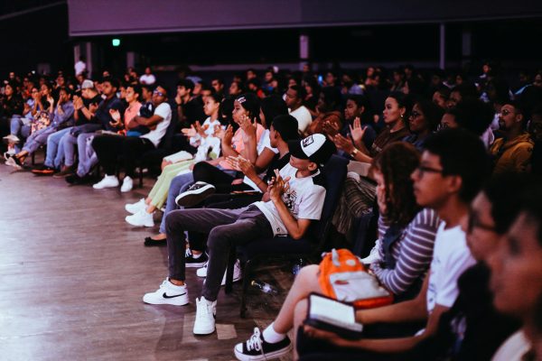 A diverse crowd seated in an auditorium applauds a live performance.