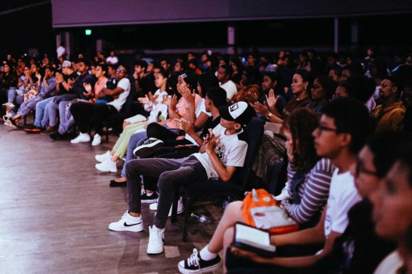 A diverse crowd seated in an auditorium applauds a live performance.