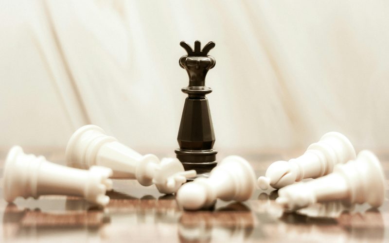 A black chess queen stands triumphant among toppled white pieces on a reflective surface.