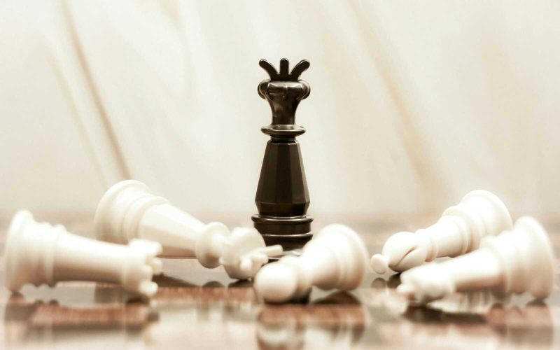 A black chess queen stands triumphant among toppled white pieces on a reflective surface.