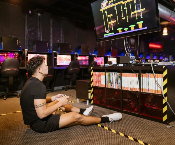 Man sitting on floor intensely playing video game at a modern arcade with vibrant computing equipment.