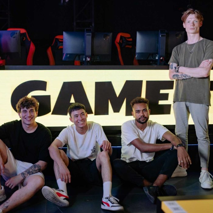 Four young men posing confidently in an eSports arena with gaming setups in the background.